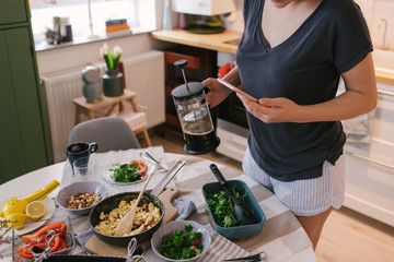 Breakfast table and woman holding smartphone in one hand navigating on social media and holding a big coffee filter glass mug with other hand, poring coffee.