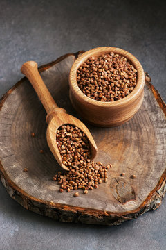 Raw buckwheat groats in a wooden scoop and bowl on a cutting board stump, dark background. Selective focus.