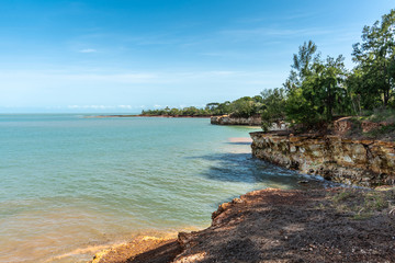 Fototapeta na wymiar Darwin Australia - February 22, 2019: Long view on East Point Shoreline shows brown, yellow and darker rocky cliffs covered by green vegetation under blue sky and greenish sea water of Beagle Gulf.