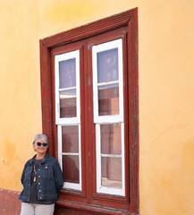 Happy elderly woman white hair smiling in front to a rustic and ancient wooden red window, spanish...