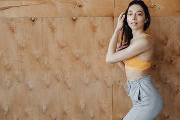 Young attractive fitness girl standing near the window on the background of a wooden wall, resting on yoga classes