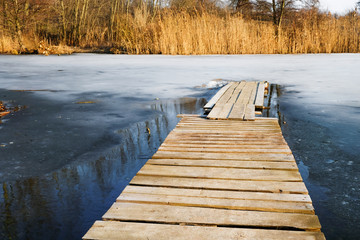 Deserted wooden bridge, melting ice and dry cane on the lake in winter in Ukraine. Self-isolation concept.