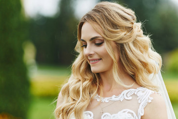 Fototapeta premium Young blonde bride with a beautiful smile and with stylish wedding hairstyle in lace white dress outdoors in the morning of the wedding day