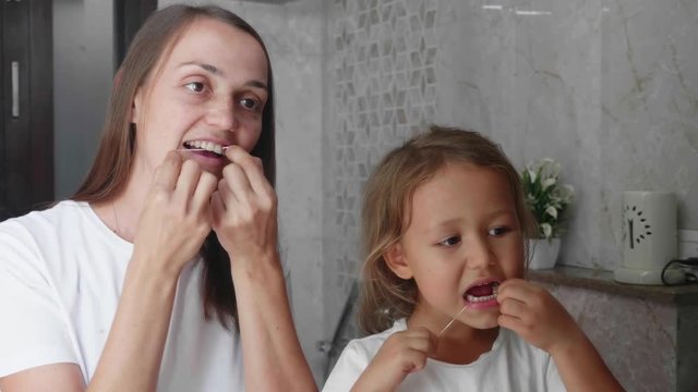 Family portrait of young mom and little cute daughter brushing their teeth with dental floss in domestic bathroom in front of the mirror.