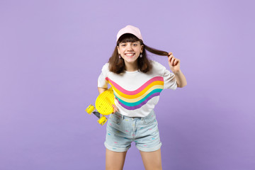 Portrait of smiling teen girl in vivid clothes with yellow skateboard, holding hair isolated on violet pastel wall background in studio. People sincere emotions, lifestyle concept. Mock up copy space.