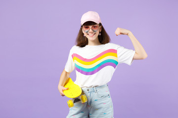 Smiling teen girl in vivid clothes, eyeglasses holding yellow skateboard, showing biceps, muscles isolated on violet pastel background. People sincere emotions, lifestyle concept. Mock up copy space.