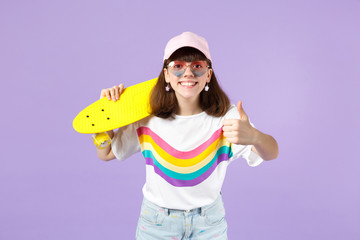 Joyful teen girl in vivid clothes, heart eyeglasses hold yellow skateboard, showing thumb up isolated on violet pastel wall background. People sincere emotions, lifestyle concept. Mock up copy space.
