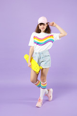 Fototapeta na wymiar Portrait of smiling teen girl in vivid clothes standing, holding yellow skateboard, rising hand isolated on violet pastel background. People sincere emotions, lifestyle concept. Mock up copy space.