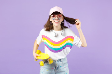 Cheerful teen girl in vivid clothes heart eyeglasses with headphones holding yellow skateboard isolated on violet pastel wall background. People sincere emotions lifestyle concept. Mock up copy space.