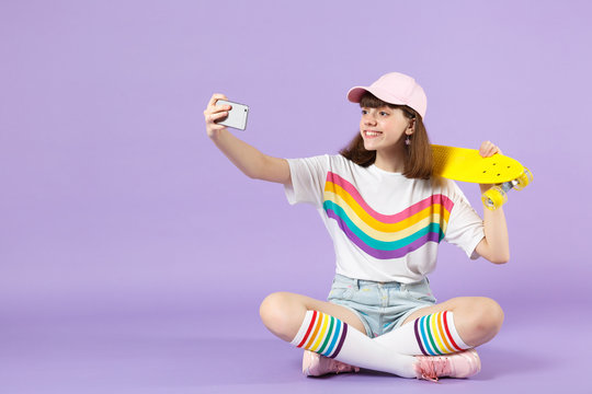 Smiling teen girl in vivid clothes holding yellow skateboard, doing selfie shot on mobile phone isolated on violet pastel background. People sincere emotions, lifestyle concept. Mock up copy space.