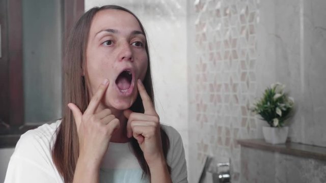 Young woman is doing facial gymnastics against wrinkles around the lips, opening her mouth and massaging her cheeks with her fingers. Portrait shot in mirror reflection.