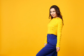 Side view of attractive young woman in sweater blue trousers standing looking camera isolated on yellow orange wall background in studio. People sincere emotions lifestyle concept. Mock up copy space.