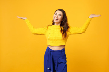 Laughing young woman in sweater, blue trousers looking camera spreading, pointing hands aside isolated on yellow orange wall background. People sincere emotions, lifestyle concept. Mock up copy space.