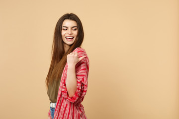 Side view of cheerful young woman in casual clothes looking, pointing thumb aside isolated on pastel beige wall background in studio. People sincere emotions, lifestyle concept. Mock up copy space.