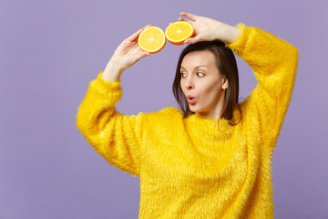 Amazed young woman in fur sweater looking aside hold in hands halfs of fresh ripe orange fruit isolated on violet pastel background. People vivid lifestyle relax vacation concept. Mock up copy space.