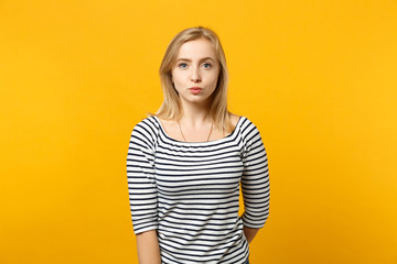 Portrait of beautiful young woman in striped clothes looking camera, blowing lips isolated on yellow orange wall background in studio. People sincere emotions, lifestyle concept. Mock up copy space.