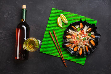 Fried rice with seafood mussels, shrimps, basil in a black plate with wine bottle, wineglass, lemon, chopsticks on green bamboo mat and stone table