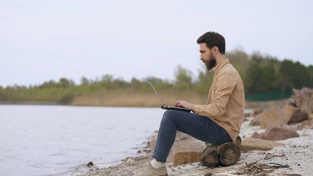 Young man working outdoors at the nature. Sitting on the coast, opening laptop. Ready to work.