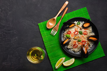 Fried rice with seafood mussels, shrimps, basil in a black plate with wineglass, lemon, wooden spoon and fork on green bamboo mat and stone table