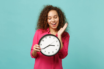 Portrait of cheerful cute african girl in pink casual clothes holding round clock isolated on blue...
