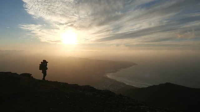 Silhouette of a woman in bicycle helmet with backpack taking pictures of an incredible sunset on the top of a Mountain above the ocean on Canary Islands. Lanzarote.