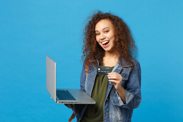 Young african american girl teen student in denim clothes, backpack work on pc, hold bank card isolated on blue wall background. Education in high school university college concept. Mock up copy space