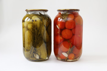 two glass jars with tomatoes and cucumbers closed with a metal lid on light background