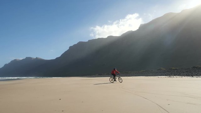 Young man rides a bicycle on a sand beach on Canary Islands towards waves of Atlantic ocean. Lanzarote