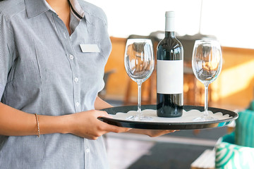 Young waitress carries a bottle of red wine with wine glasses on a tray to the customer of the hotel restaurant. The concept of service.