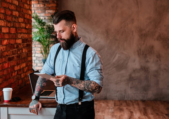 Serious bearded man is leaning on the table. He is wearing shirt and suspender. He has tattoes on...