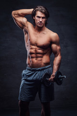 Attractive shirtless bodybuilder is posing with dumbbell. There is dark background.
