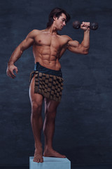 Obraz premium Beautiful strong bodybuilder is posing on white pedestal with naked torso, dumbbell and gladiator's bandage.