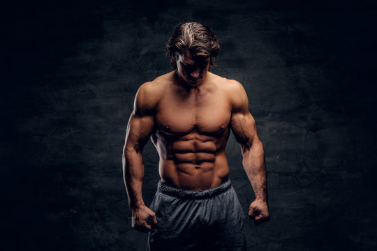 Young attractive bodybuilder with naked torso is posing for photographer on grey background.