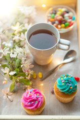 Bright cakes. Cupcakes with a cup of coffee or tea and color dots on a tray with a flowering branch and a luminous garland on a white background. Morning mood, spring background