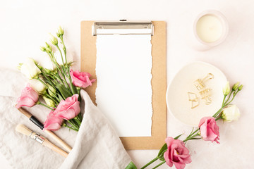Top view with blank paper clipboard, paint brushes, napkin, rose and white blooming flowers, candle and office accessories. Flat lay with copy space. Home office desk in rose, white, gold colors