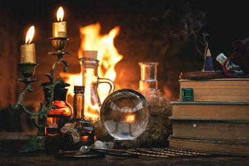Crystal ball, magic book, magic potion and other wizard accessories on a table on a burning fire background.