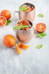 Blood orange Moscow mule alcohol cocktail with fresh mint leaves and ice in copper mugs on a gray concrete background.
