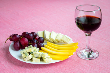 Glass of red wine and tree kinds of sliced cheese and sweet red grapes on white plate. Cheeses covered with edible white and blue mold. Still life on a pink tablecloth.
