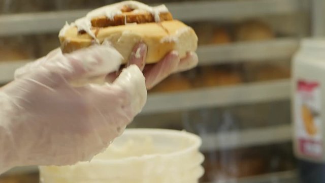 With Gloved Hand Woman Puts Icing On Several Cinnamon Roles. Woman ices cinnamon rolls at professional bakery.
