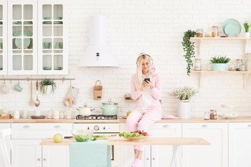 Fototapeta na wymiar Young Woman in cozy pajamas and headphones cooking in the kitchen and listening fun music. Healthy Food - Vegetable Salad. Diet. Dieting Concept. Healthy Lifestyle. Cooking At Home. 