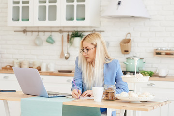 Freelance concept. Beautiful business woman in casual clothes and glasses is examining documents and smiling while working with a laptop in kitchen. Working at home