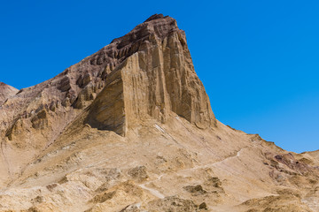 A hiking trail traverses the base of a high desert mountain peak in a vast colorful desert landscape -  Manly Beacon in Death Valley National Park
