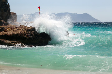 Fototapeta premium Waves coming from the sea are blasting on the rocks. Water color is turquoise and a red yellow flag is on the rock. Windy and stormy weather at Mediterranean Sea. Buyukcakil Beach, Antalya Turkey