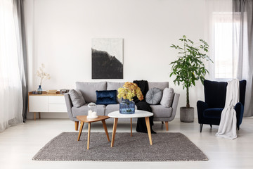 Flowers in blue glass vase and two white candles on wooden coffee tables in grey elegant living room with comfortable sofa