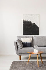 Abstract black and white painting on empty white wall with wooden coffee table with candles and grey sofa with pillow