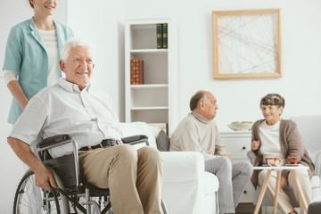 Fototapeta premium Smiling nurse supporting happy elderly man in the wheelchair near senior people