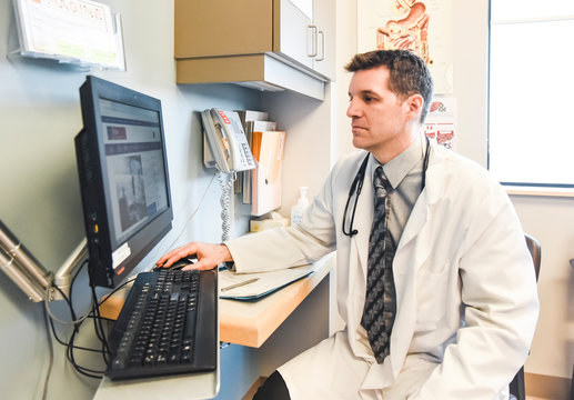 Doctor in a white coat using a computer at a desk in a clinic room.