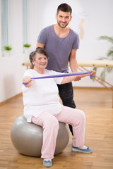 Elderly lady siting on the exercise ball during physiotherapy with her young instructor