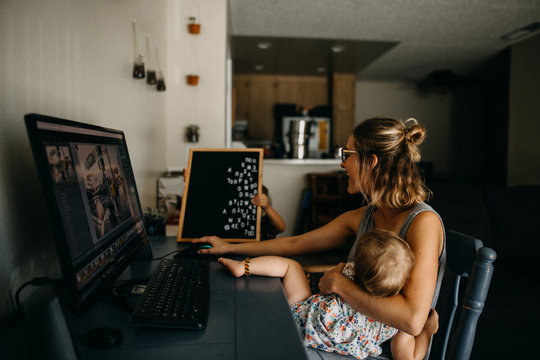 Woman holding her daughter while working on laptop at home