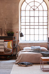 Lamp between bed and basket with pillows in industrial bedroom interior with window and plant. Real photo